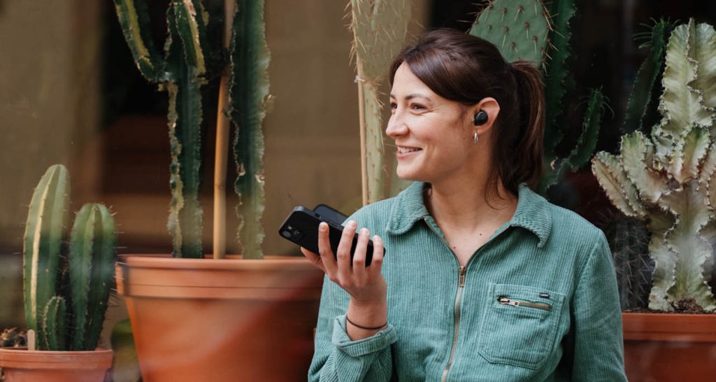 Een glimlachende vrouw draagt een in-ear koptelefoon en houdt een smartphone in haar hand.