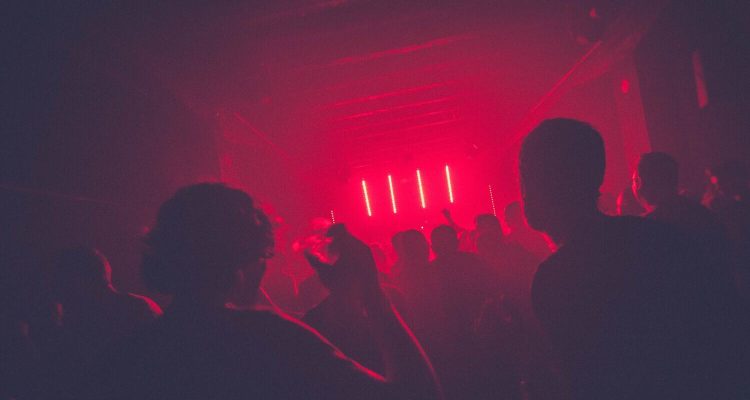 Menschen tanzen in einem dunklen Club mit rot schimmerndem Licht im Hintergrund.