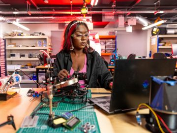 Frau arbeitet in einer Werkstatt an technischen Bauteilen und einem Laptop