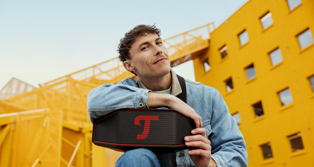 Een man poseert met de ROCKSTER CROSS 2 van Teufel voor een geel gebouw.