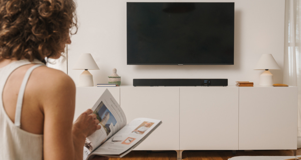 CINEBAR LUX Surround Ambition "5.1-Set" Vrouw zit in woonkamer. Voor haar een tv-meubel met daarop de CINEBAR 22 en een tv die aan de wand is gemonteerd