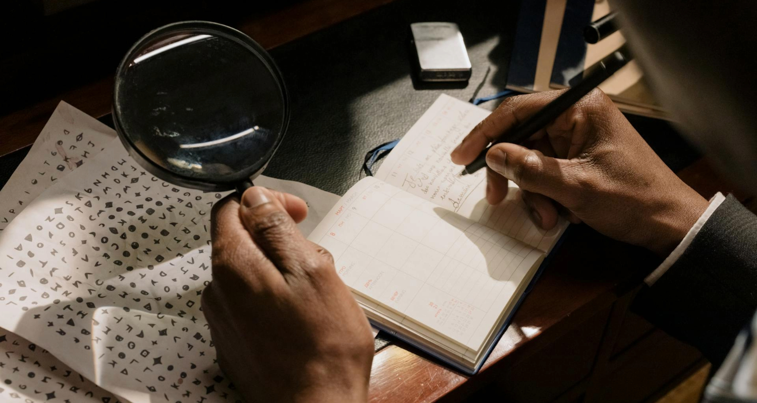 Person untersucht ein Blatt mit Symbolen durch eine Lupe und macht Notizen in ein geöffnetes Notizbuch.
